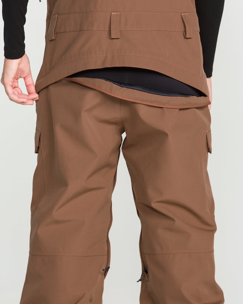 A person is shown wearing the Volcom Women’s Creston 3D Stretch Bib in brown, paired with a matching brown jacket. The jacket’s back flap is lifted to reveal a layer of recycled stretch oxford underneath. The image highlights the waist and lower back area, with hands visible at the sides.