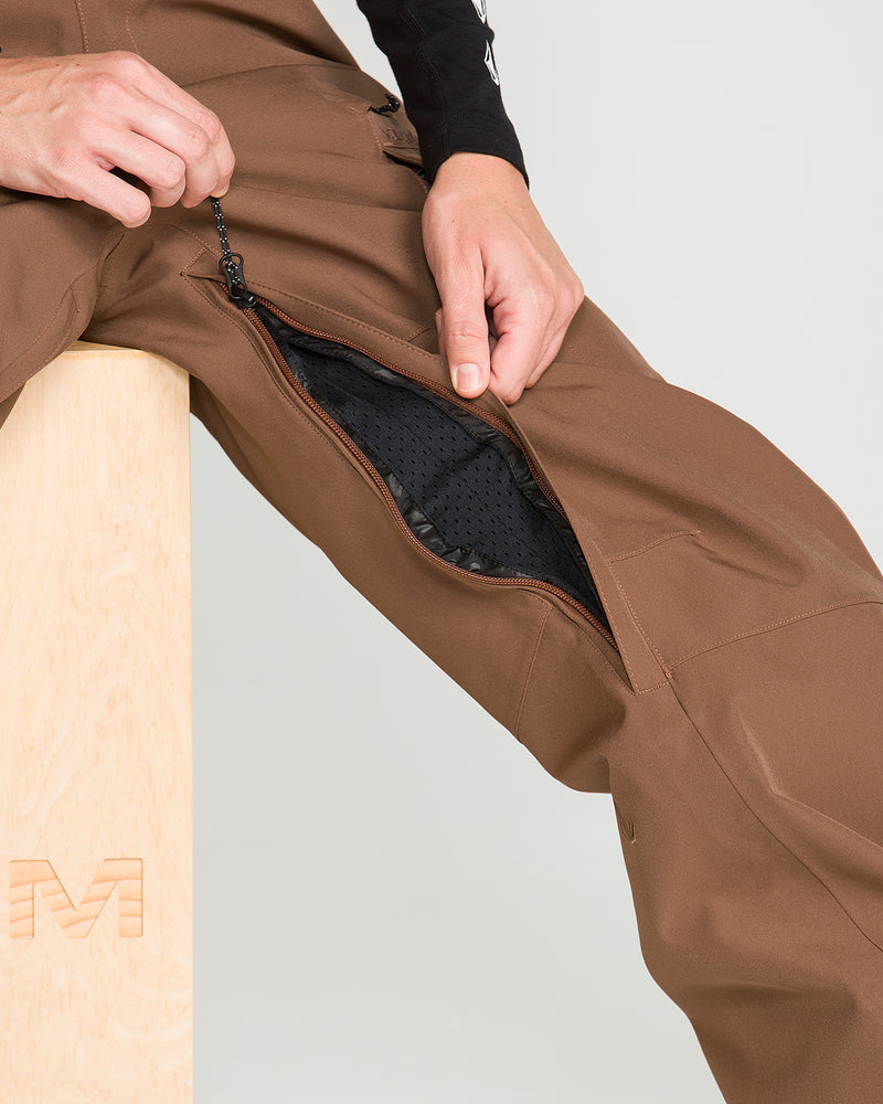 A person sitting on a wooden block unzips a vent on the side of their Volcom Women’s Creston 3D Stretch Bib, made from recycled stretch oxford in brown, revealing black mesh fabric inside.