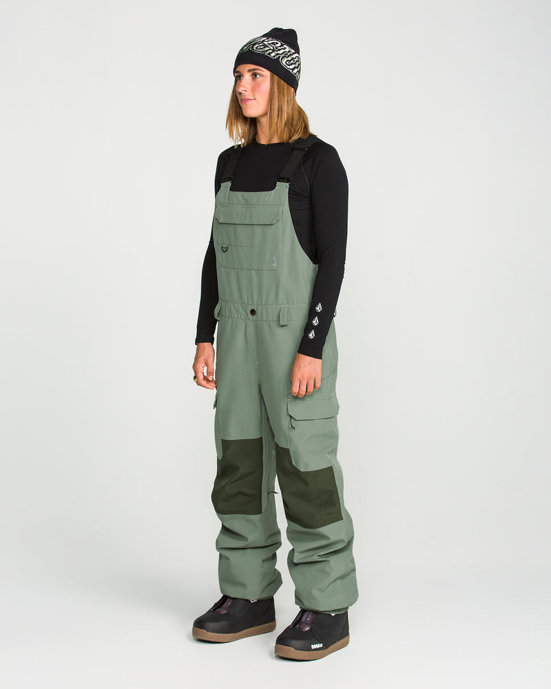 A person wearing a black long-sleeve shirt, Volcom Women’s Creston 3D Stretch Bib overalls in olive green with dark knee patches, dark snow boots, and a black beanie stands facing left against a plain white background.