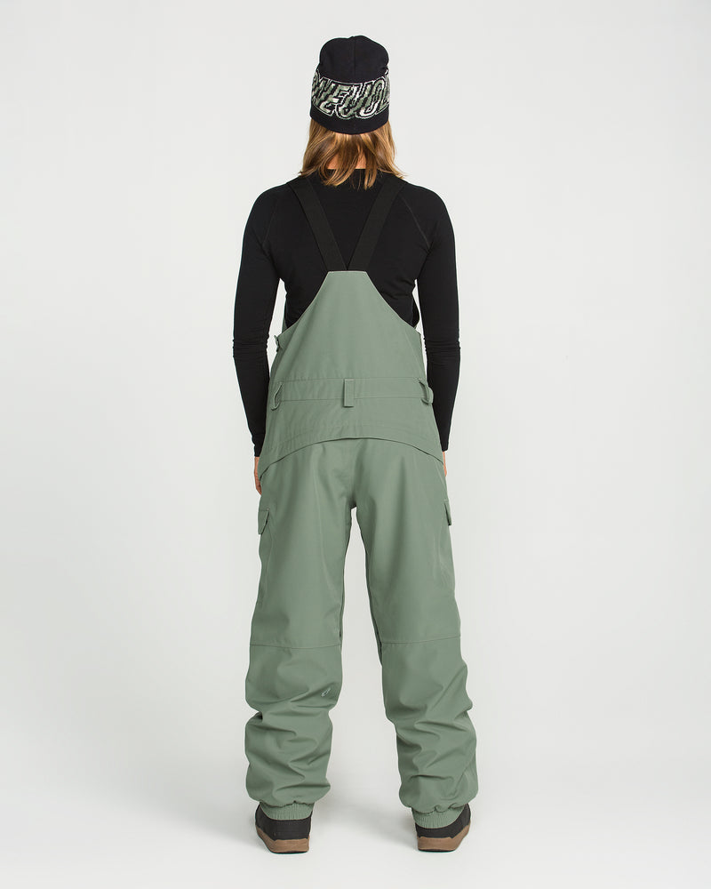 A person stands with their back to the camera, wearing Volcom Women’s Creston 3D Stretch Bib snow pants in sage green—crafted from recycled stretch oxford fabric with fully taped seams—paired with a black long-sleeve shirt, brown boots, and a black beanie featuring a white pattern, set against a plain white background.