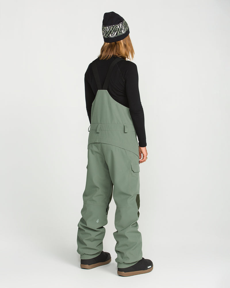 A person stands with their back to the camera, dressed in a black long-sleeve shirt and the Volcom Women’s Creston 3D Stretch Bib in sage green, made from recycled stretch oxford. The look is completed with black boots and a black beanie featuring white lettering, set against a plain white background.