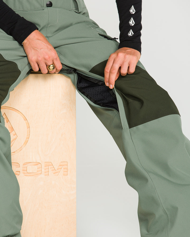 A person opens a zipper vent on the Women’s Creston 3D Stretch Bib by Volcom—green and black pants made from recycled stretch oxford material—while sitting on a wooden box. Featuring mesh lining, cargo-style pockets, and fully taped seams, only the lower torso and hands are visible.