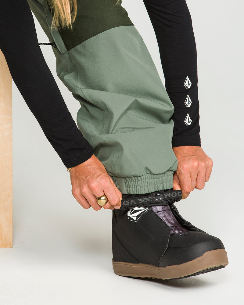 A person wearing Volcom Women’s Creston 3D Stretch Bib pants in green—crafted from recycled stretch oxford fabric with fully taped seams—adjusts the cuff over a black snow boot featuring the Volcom logo. Only their lower body and hands are visible, paired with a black long-sleeve shirt.