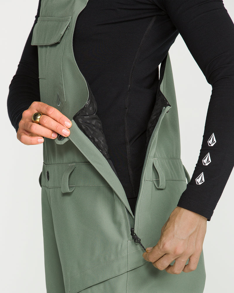 A person wearing a black long-sleeve shirt and Volcom Women’s Creston 3D Stretch Bib overalls in green unzips the side of the overalls, revealing a black patterned inner lining. Only the torso and hands are visible.