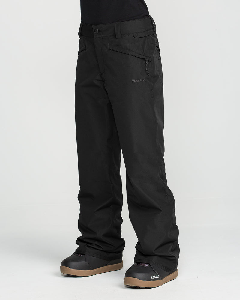 A person is shown from the waist down wearing black Volcom Women’s Coldfront Insulated Baggy Pants and black snow boots, standing against a plain white background.