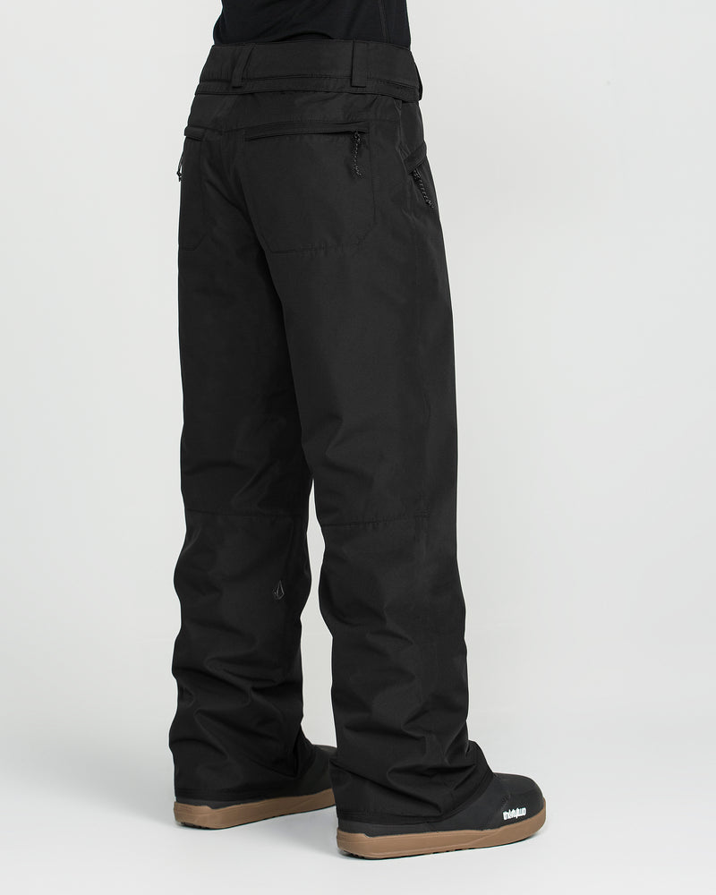 A person wearing Volcom Women’s Coldfront Insulated Baggy Pants in black, featuring V-Science 2-Layer construction, paired with black snow boots with brown soles, standing against a plain white background.