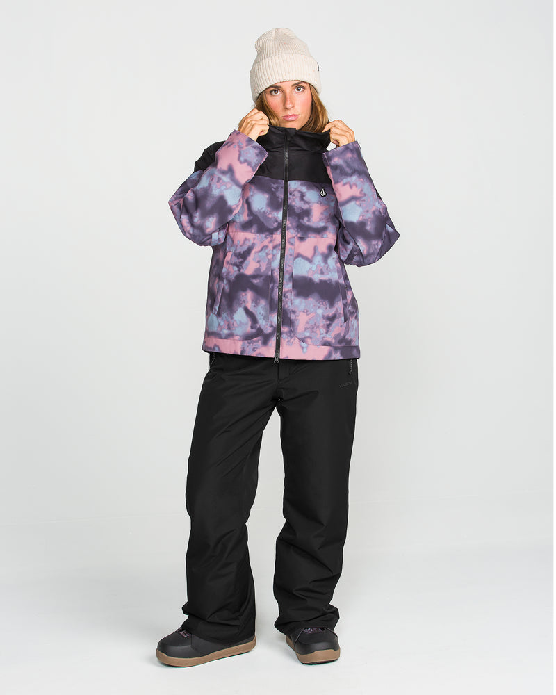 A person wearing a beige beanie, a purple and black tie-dye V-Science 2-Layer jacket with recycled insulation, Volcom Women’s Coldfront Insulated Baggy Pants, and dark boots stands against a plain light background, looking forward with hands holding the jacket collar.