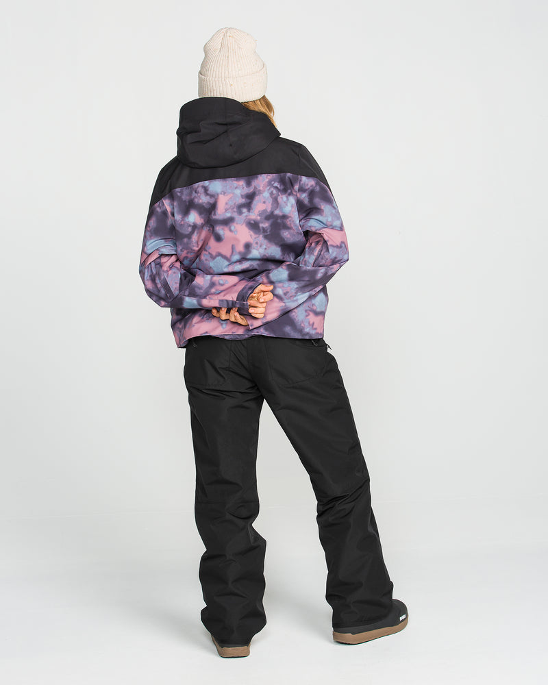 A person stands with their back to the camera, wearing a beige beanie and a purple and black tie-dye jacket featuring V-Science 2-Layer technology. They are also wearing Volcom Women’s Coldfront Insulated Baggy Pants in black. Their hands are clasped behind them as they stand against a plain white background.