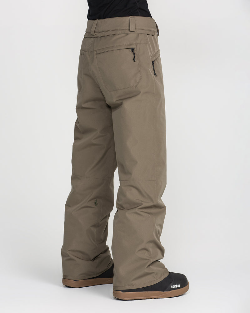 A person is wearing Volcom Women’s Coldfront Insulated Baggy Pants in khaki along with black winter boots featuring brown soles, standing on a plain white background and facing slightly away from the camera.