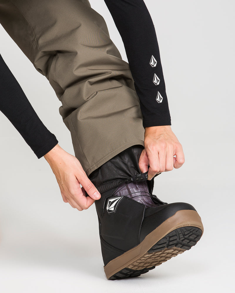 Wearing the Volcom Women’s Coldfront Insulated Baggy Pants in brown, a person adjusts the cuff over a black snow boot with a brown sole, pulling down the pant leg to reveal visible Volcom logos on both the sleeve and the boot.