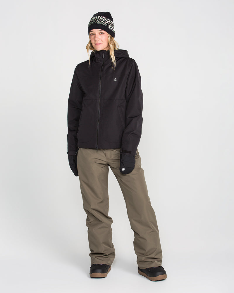 A person stands against a plain white background wearing black winter boots, black gloves, a black hooded jacket, a black beanie, and Volcom Women’s Coldfront Insulated Baggy Pants in olive green.