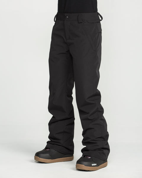 A person is wearing Volcom Women’s Stoned Insulated Pants in black along with black snow boots featuring brown soles, standing against a plain white background. Only the lower half of the body is visible.
