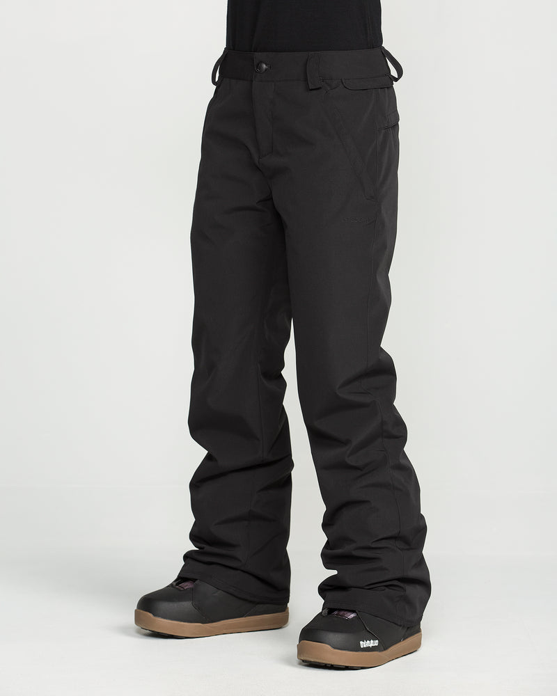 A person is wearing Volcom Women’s Stoned Insulated Pants in black along with black snow boots featuring brown soles, standing against a plain white background. Only the lower half of the body is visible.
