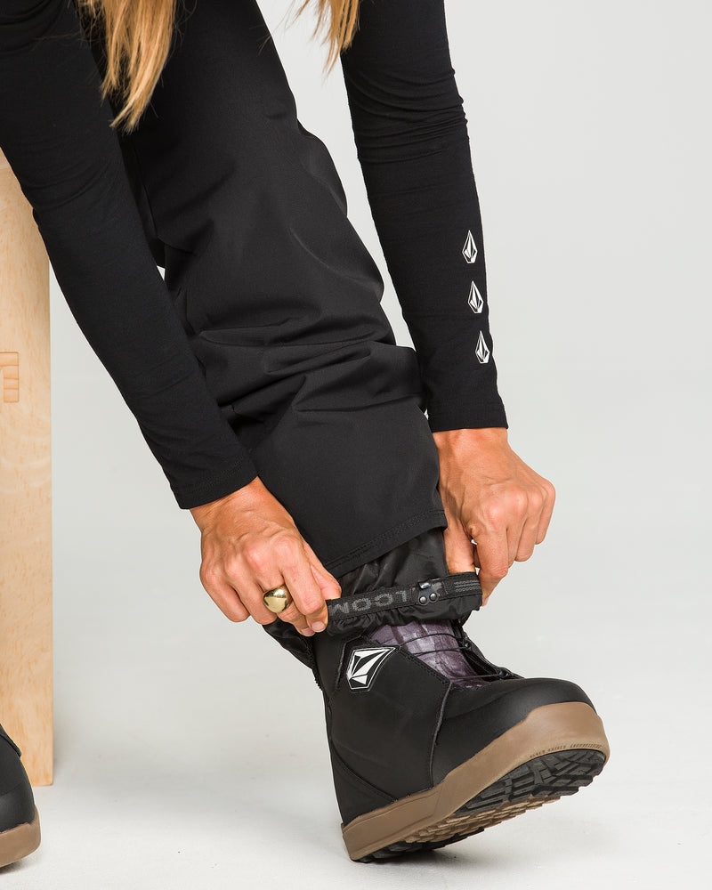 A person in black clothing adjusts the cuff of their Volcom Women’s Stoned Insulated Pants—featuring a 10K waterproof rating—over a black snow boot with a brown sole. Both the boot and pant leg display a geometric Volcom logo. Only the person’s hands and lower body are visible.