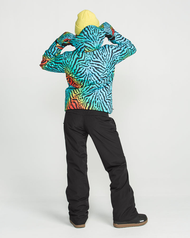 A person wearing a yellow beanie, a colorful zebra-striped jacket, and Volcom Women’s Stoned Insulated Pants in black stands facing away, adjusting their hood against a plain white background.