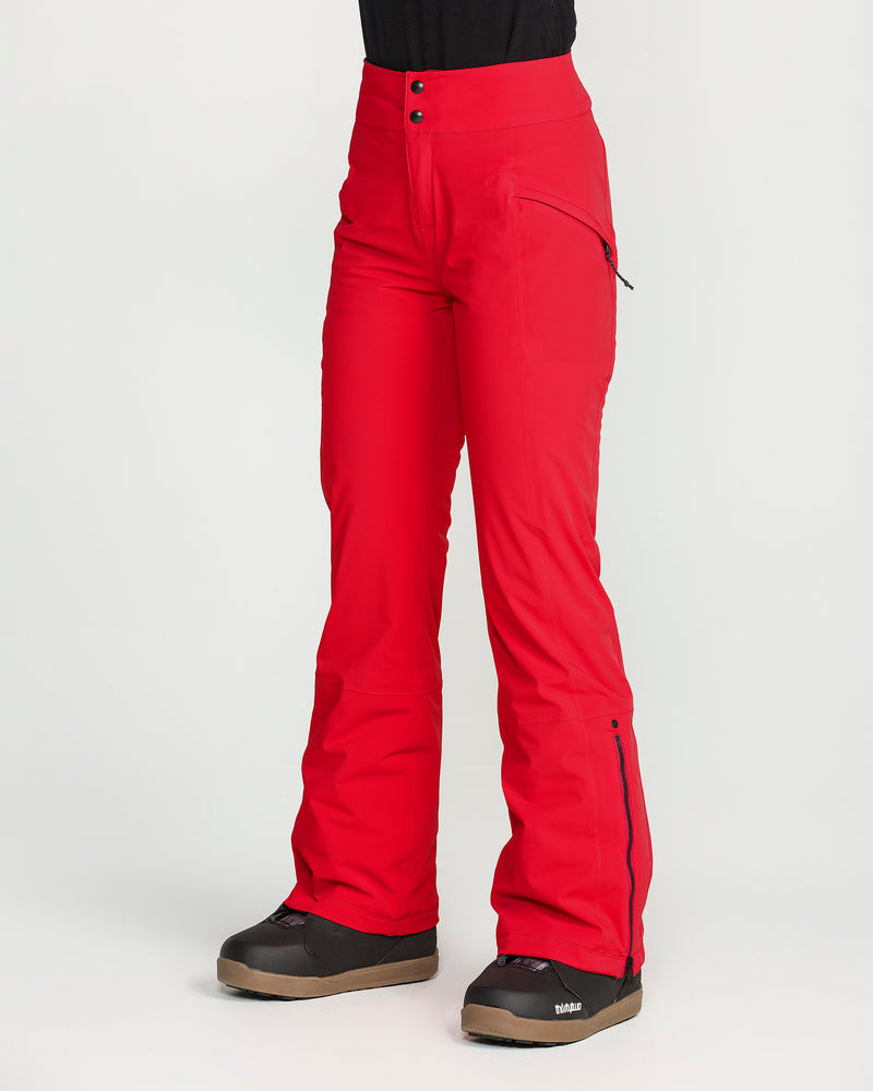 A person is wearing bright red Volcom Women’s Alpine All Day Insulated Pants and black snow boots, standing against a plain white background. These high-rise fit pants feature zippered pockets, flared legs, and two visible buttons at the waist, and are crafted with 4-way ultra stretch fabric for all-day comfort on or off the slopes.