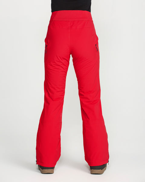 A person is seen from the back, standing on a white background, wearing bright red Volcom Women’s Alpine All Day Insulated Pants and brown boots. These pants offer 4-way ultra stretch fabric, zippered side pockets, and a high-rise fit with a comfortable waistband.