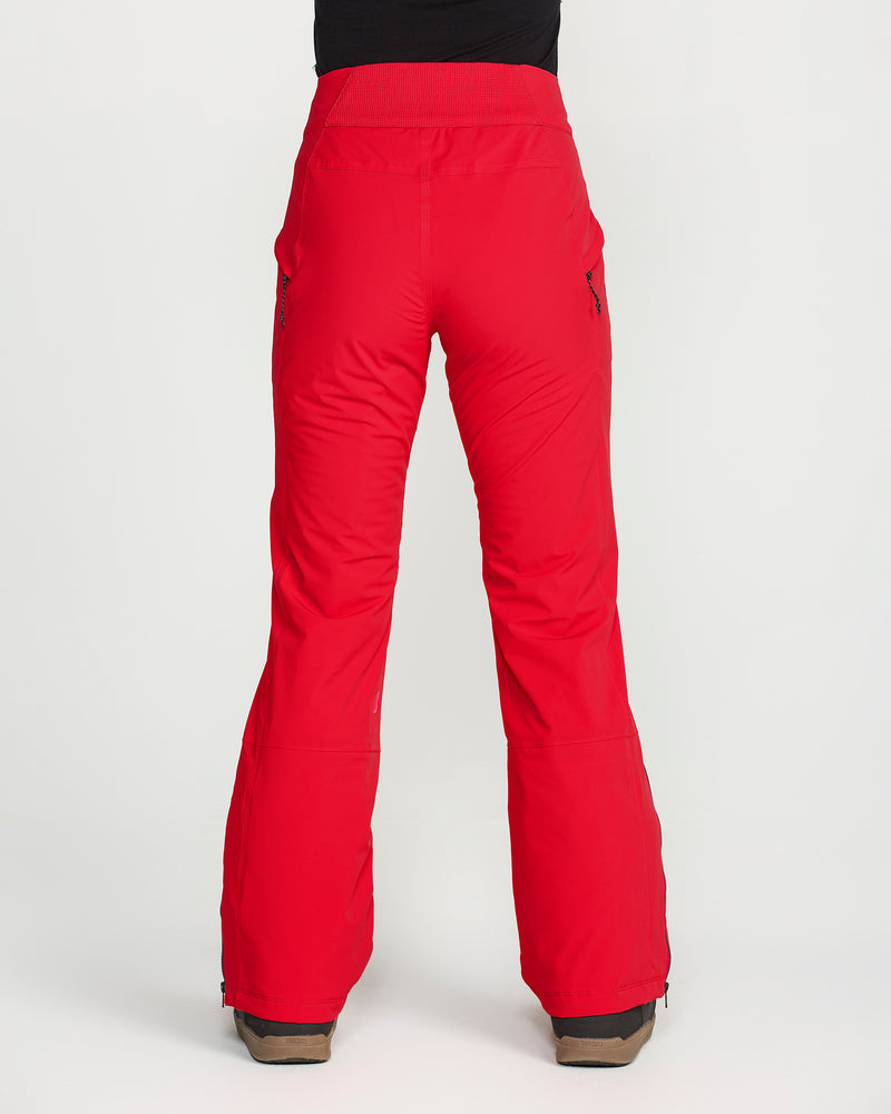A person is seen from the back, standing on a white background, wearing bright red Volcom Women’s Alpine All Day Insulated Pants and brown boots. These pants offer 4-way ultra stretch fabric, zippered side pockets, and a high-rise fit with a comfortable waistband.