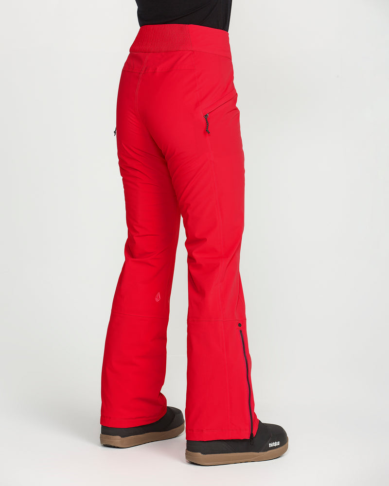 A person is shown standing sideways, wearing the Volcom Women’s Alpine All Day Insulated Pants in bright red. These snow pants feature a high-rise fit, zippered pockets, and 4-way ultra stretch for maximum comfort. The pants have a slight flare at the bottom and are paired with black winter boots, all set against a white background.