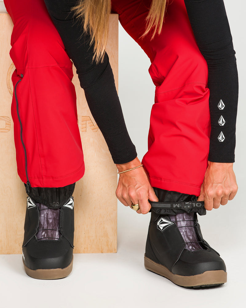 Wearing the Volcom Women’s Alpine All Day Insulated Pants in red with a high-rise fit and a black long-sleeve shirt, she sits on a wooden box tightening the strap on her black snowboard boot.