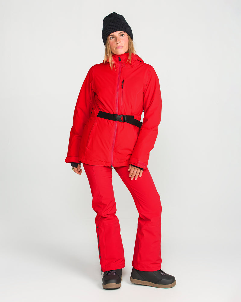 A person wears a red ski jacket and the Volcom Women’s Alpine All Day Insulated Pants, which offer 4-way ultra stretch and a comfortable high-rise fit. The outfit is completed with black boots and a black beanie, set against a plain white background.