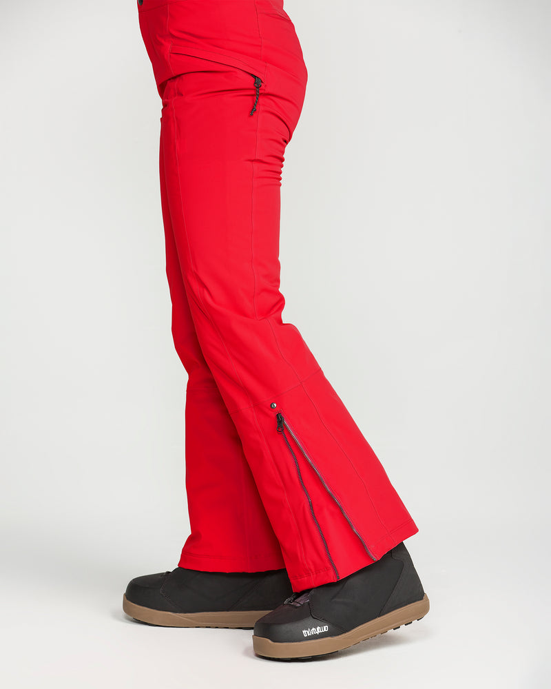 A person wearing Volcom Women’s Alpine All Day Insulated Pants in bright red, featuring 4-way ultra stretch and zippered side vents, paired with black snow boots. Posed sideways against a plain white background, only the lower body is visible.