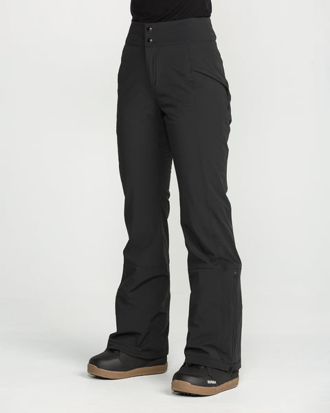 A person is shown wearing Volcom Women’s Alpine All Day Insulated Pants in black, featuring a high-rise fit and flared legs, paired with black snow boots with tan soles. The individual is standing on a white background, and only the lower body is visible.