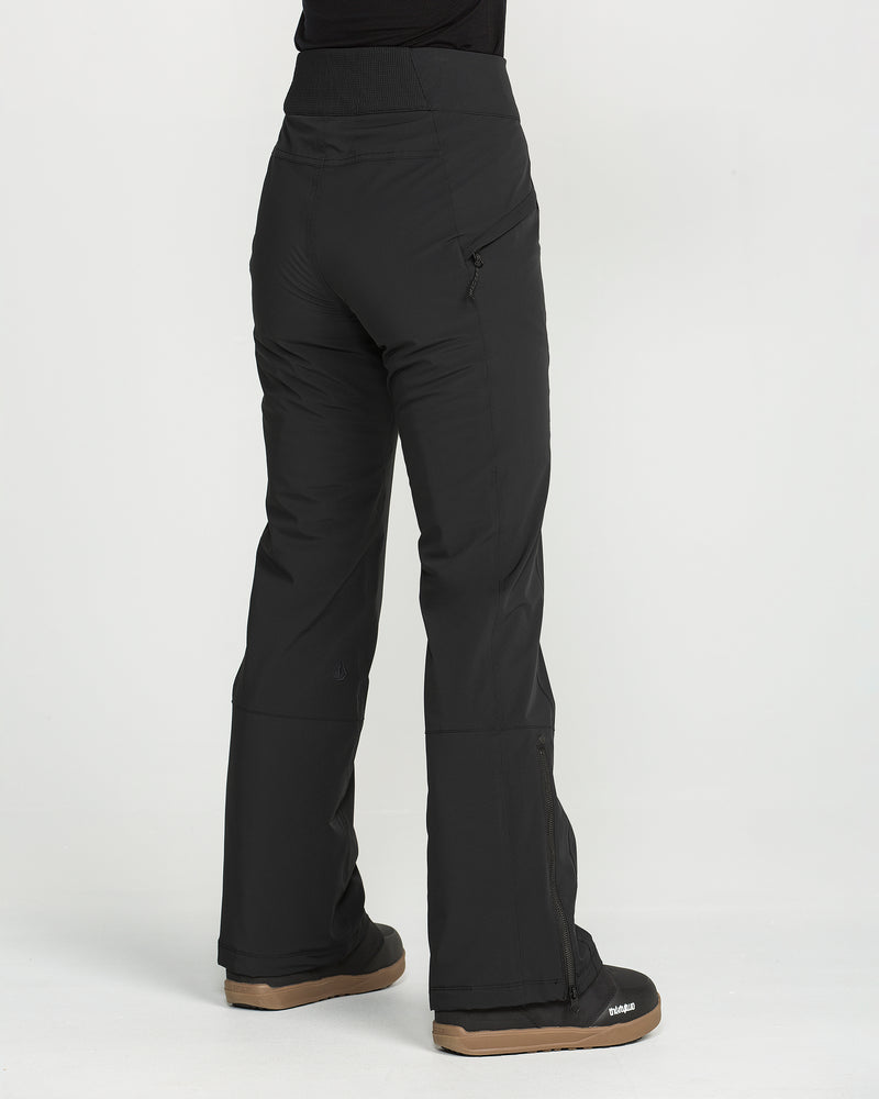 A person is wearing the Volcom Women’s Alpine All Day Insulated Pants in black and black boots, standing on a white background and facing away from the camera. These pants feature a slim, high-rise fit with a zipped pocket and boot-cut hem.