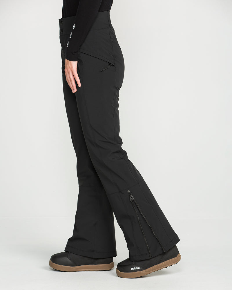 A person wearing black Volcom Women’s Alpine All Day Insulated Pants and black boots stands sideways against a plain white background. The pants, designed with a high-rise fit, feature zippers at the sides and a fitted waist.