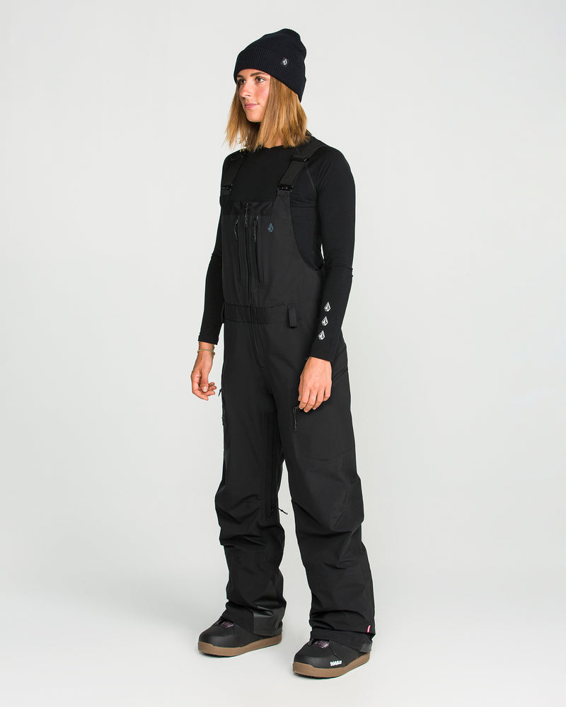 A person wearing a black beanie, long-sleeve shirt, and Volcom Women’s Vs 3L Stretch Gore Bib pants stands against a plain white background.
