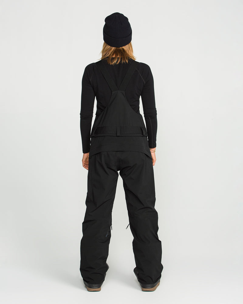 A person is seen from behind against a plain white background wearing the Volcom Women’s Vs 3L Stretch Gore Bib in black, paired with a black long-sleeve shirt and a black beanie.