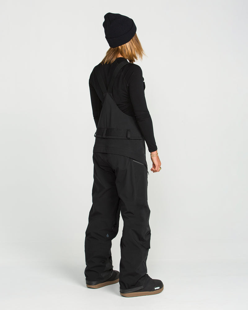 A person wearing a black beanie, long-sleeve shirt, and Volcom Women’s Vs 3L Stretch Gore Bib ski pants stands turned away from the camera against a plain white background.