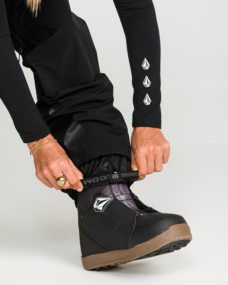 A person wearing Volcom Women’s Vs 3L Stretch Gore Bib ski pants and black snow boots adjusts a strap around the boot, their hands adorned with a ring and bracelets. The Volcom logo is visible on both the boot and sleeve, emphasizing the PFAS-free design.