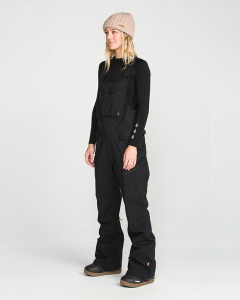 A person stands in a studio on a white background, looking slightly to the side with a neutral expression, wearing a beige knit beanie, a black long-sleeve shirt, and the Volcom Women’s Ash Stretch Gore Bib.