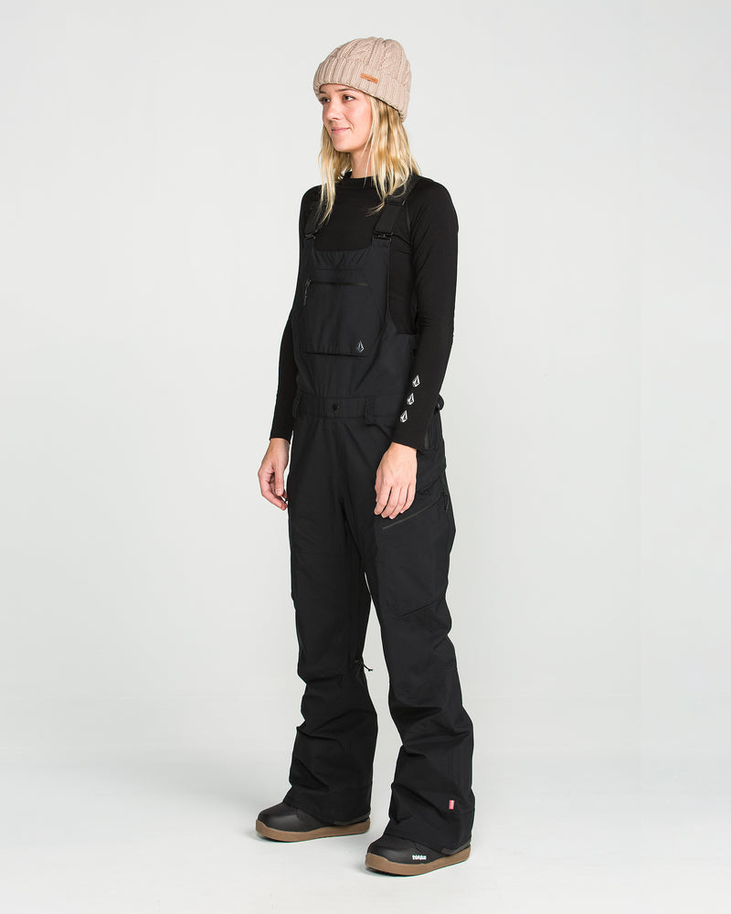 A person stands in a studio on a white background, looking slightly to the side with a neutral expression, wearing a beige knit beanie, a black long-sleeve shirt, and the Volcom Women’s Ash Stretch Gore Bib.