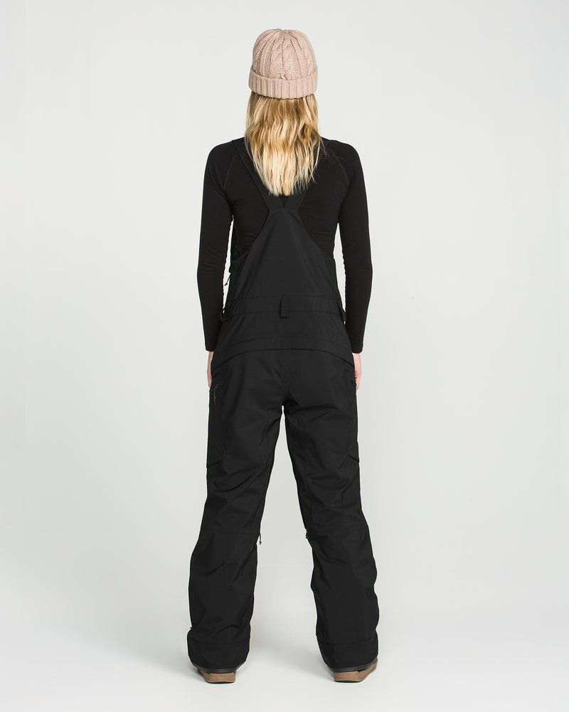 A person with long blonde hair, wearing a beige knit beanie, a black long-sleeve top, and Volcom Women’s Ash Stretch Gore Bib snow pants stands facing away against a plain white background.