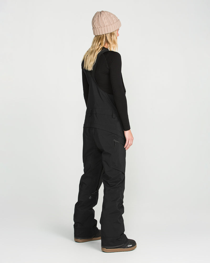 A person is shown from behind on a plain white background, wearing the Volcom Women’s Ash Stretch Gore Bib snow pants and a black long-sleeve shirt, paired with a beige knit beanie.