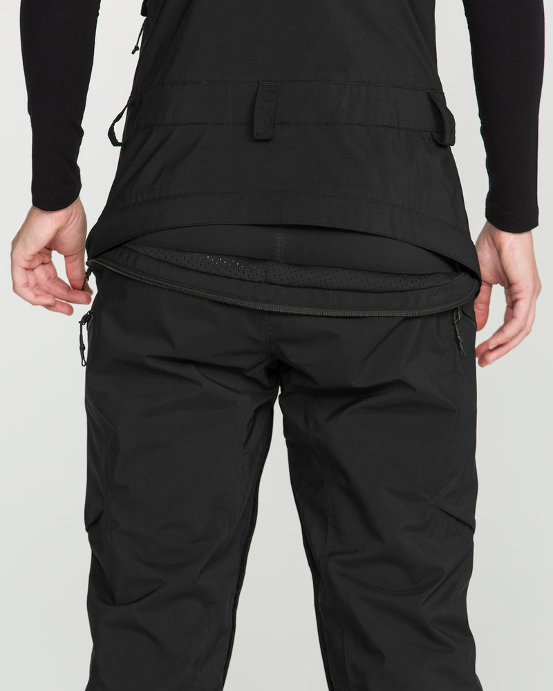 A person wearing Volcom’s Women’s Ash Stretch Gore Bib snow pants and a black jacket, with the back of the jacket unzipped to reveal the bib’s inner waistband. The image focuses on their lower back and upper legs.