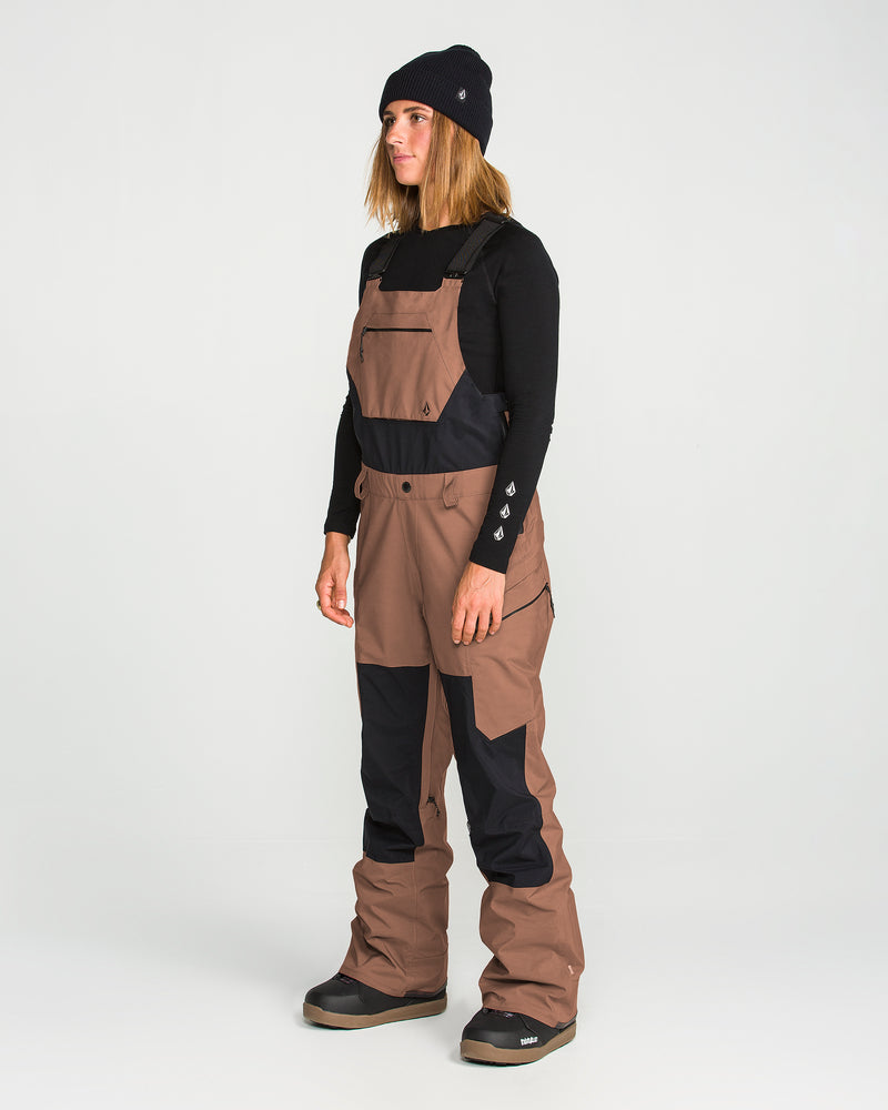 A woman wearing the Volcom Women’s Ash Stretch Gore Bib in brown and black GORE-TEX, paired with a black long-sleeve shirt, black beanie, and snow boots, stands against a plain white background.