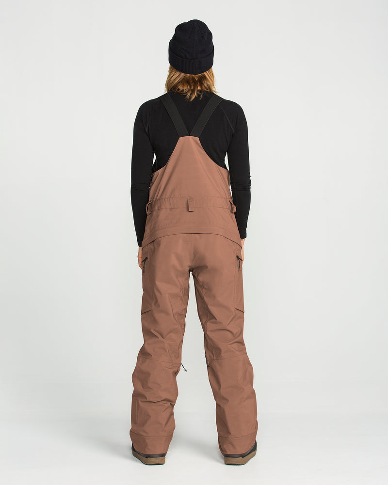 A person stands facing away, wearing a black beanie, a black long-sleeve shirt, and loose brown Volcom Women’s Ash Stretch Gore Bib overalls with straps. The background is plain white.