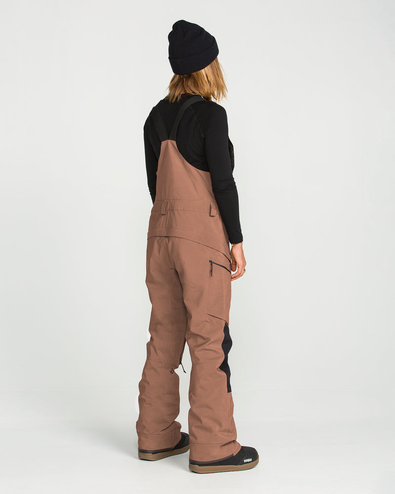 A person is shown from behind in a studio setting wearing a black beanie, black long-sleeve shirt, and Volcom Women’s Ash Stretch Gore Bib overalls in brown with black patches.