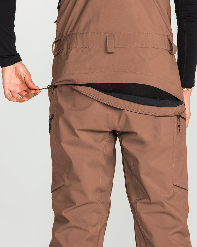 A person wearing Volcom’s Women’s Ash Stretch Gore Bib in brown unzips a vent at the back of the jacket, revealing a black inner layer. The photo, focused on the lower back and hand against a light background, subtly highlights technical features such as the waterproof GORE-TEX bib details designed to enhance performance in changing conditions.
