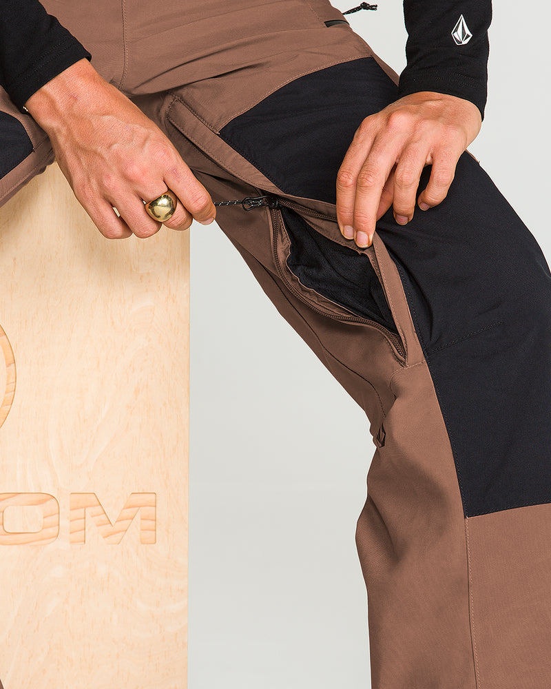 A person unzips a pocket on the thigh of Volcom Women’s Ash Stretch Gore Bib waterproof pants in a brown and black colorway while sitting on a light wooden box. Only the hands and part of the legs are visible.
