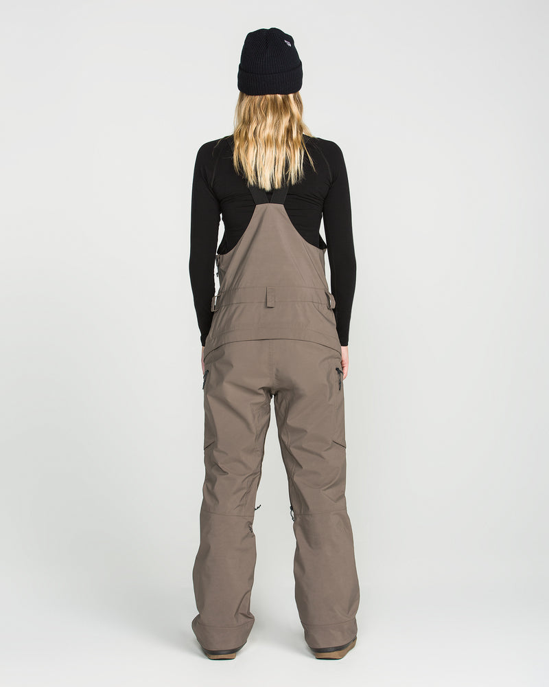 A person wearing a black beanie, black long-sleeve shirt, and the Volcom Women’s Ash Stretch Gore Bib in taupe stands facing away from the camera against a plain white background.