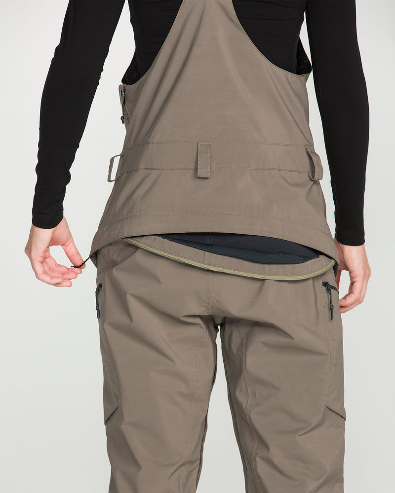 A person is seen from the back, wearing the Volcom Women’s Ash Stretch Gore Bib in taupe, styled with a back zipper reminiscent of women’s snow pants. They are dressed in black underneath and are lifting their jacket to reveal the zipper, with hands on either side.