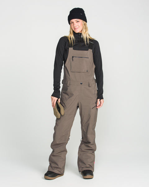 A person is wearing the Volcom Women’s Ash Stretch Gore Bib in brown, paired with a black beanie and black long-sleeve shirt, while holding gloves and smiling at the camera against a plain light background.