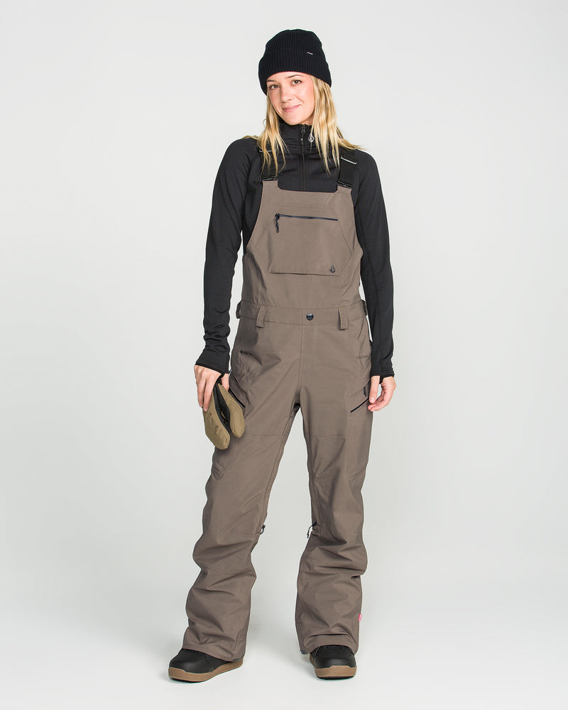A person is wearing the Volcom Women’s Ash Stretch Gore Bib in brown, paired with a black beanie and black long-sleeve shirt, while holding gloves and smiling at the camera against a plain light background.