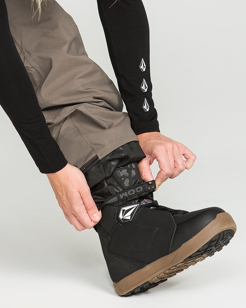 A person tucks the Volcom Women’s Ash Stretch Gore Bib snow pants into black snowboarding boots, securing the gaiter. Their hand adjusts the boot strap. The tan waterproof ski bib pants complement the boots’ brown soles. The background is plain light gray.