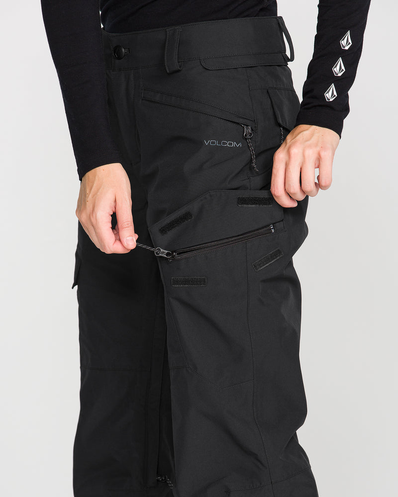 A person unzips a zipper on the thigh of black Volcom Women’s Stonelark Gore-Tex Pants, revealing a zip-off feature. The person is wearing a black long-sleeve shirt with white logos on the sleeve.