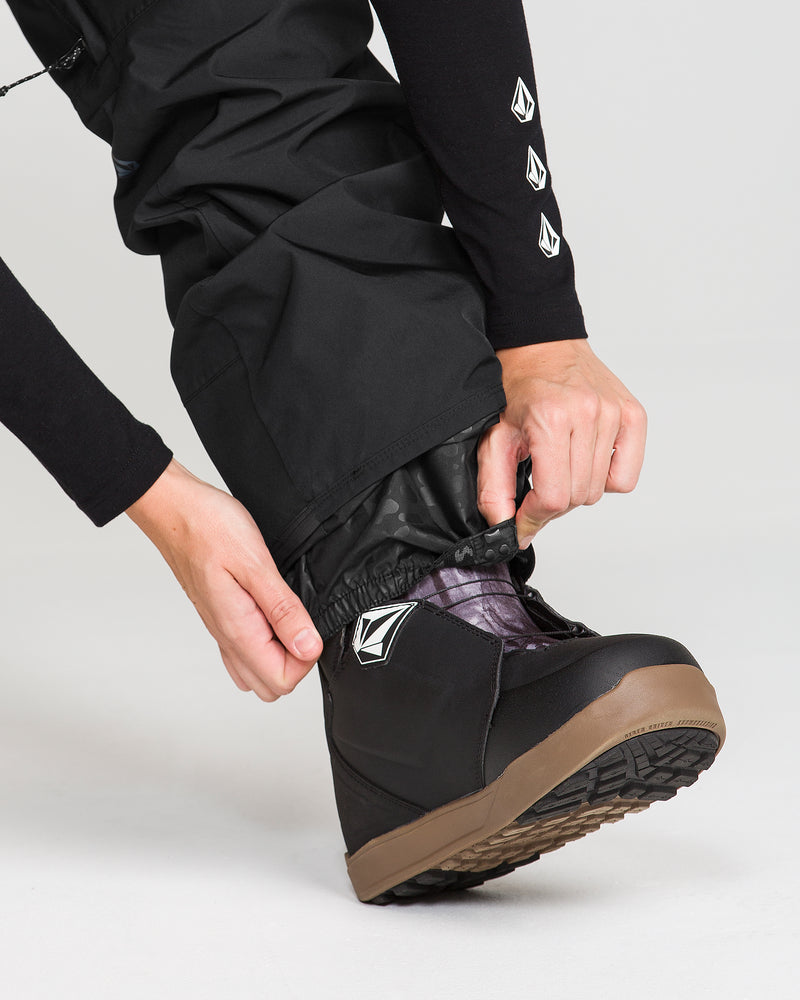 A person adjusts the inner gaiter of their black Volcom Women’s Stonelark GORE-TEX Pants over a black snow boot with a brown sole. They are wearing a black long-sleeve shirt featuring a white logo on the sleeve.